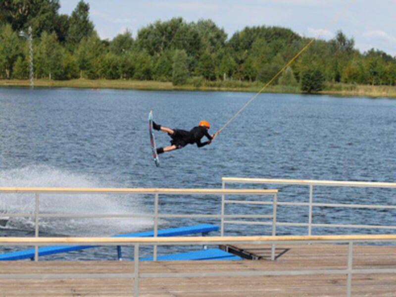 Un wakeboarder en combinaison noire et casque orange réalise un saut acrobatique au-dessus d'un ponton en bois sur un lac.
