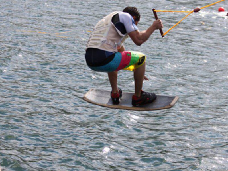 Un homme en short coloré et gilet de protection est en équilibre sur un wakeboard, tenant fermement la poignée du câble.