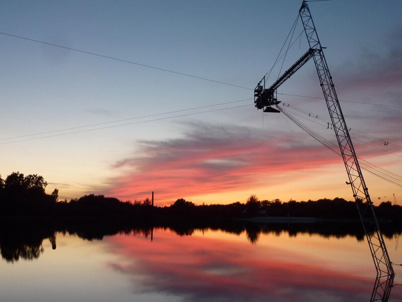 Câble et structure de wakeboard au-dessus d'un lac calme au coucher du soleil avec un ciel aux teintes roses et orangées.