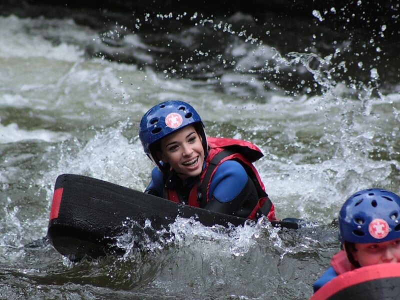 Jeune femme souriante en hydrospeed dans une rivière en eau vive, portant casque bleu et gilet de sauvetage rouge.