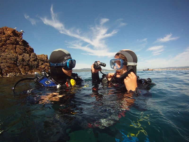 Deux plongeurs en combinaison noire s'apprêtant à plonger dans une mer calme près des rochers.