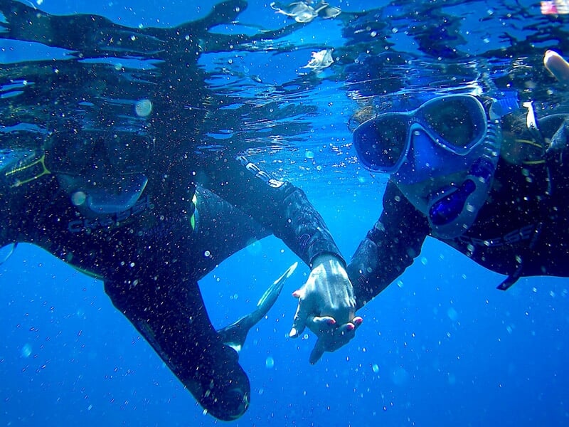 Deux plongeurs sous l'eau se tenant la main, entourés de bulles et d'une eau bleue intense.
