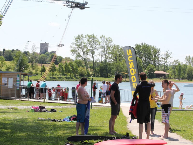 Groupe de personnes en maillots et combinaisons près d’un lac, avec un téléski nautique et un drapeau jaune sur pelouse ensoleillée.