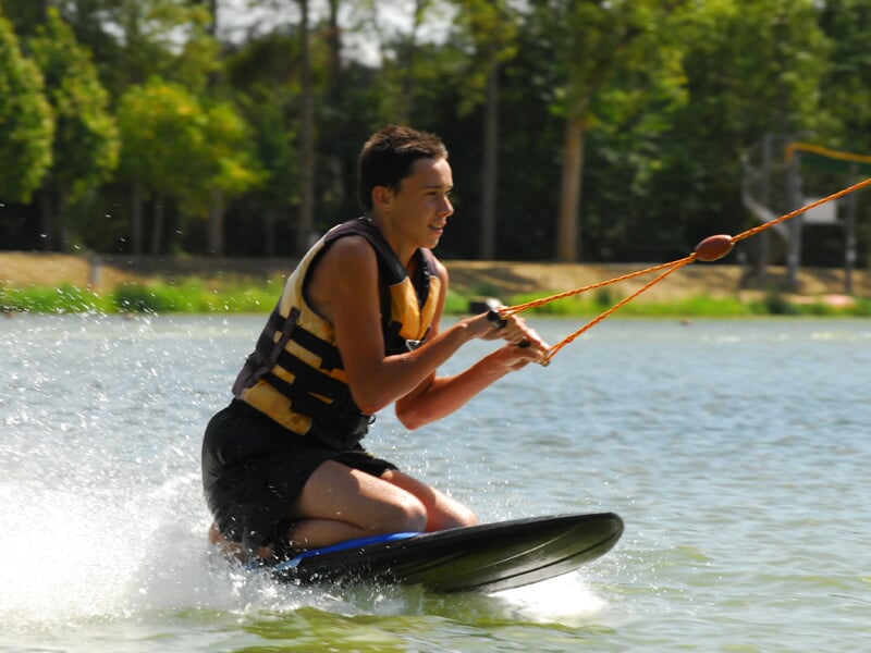 Jeune homme en gilet de sauvetage jaune et noir, agenouillé sur un kneeboard tiré par un câble sur un lac.