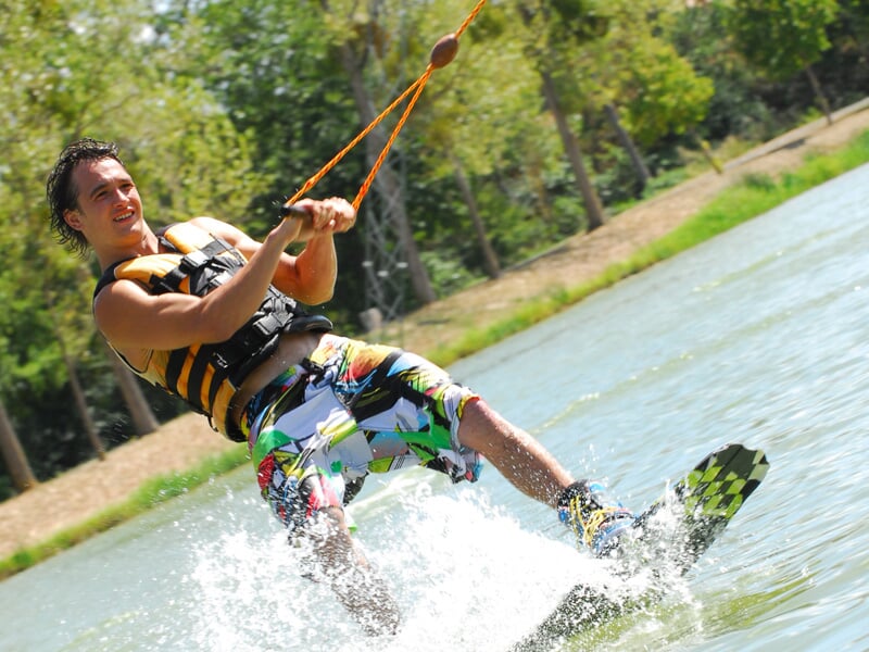 Wakeboarder souriant en short à motifs et gilet jaune et noir, éclaboussant l’eau tout en tenant une corde.