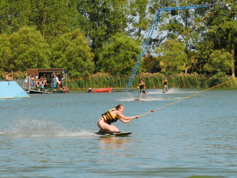 Trois wakeboarders tractés par câble sur un lac, avec une zone d’attente flottante et un module bleu à gauche.