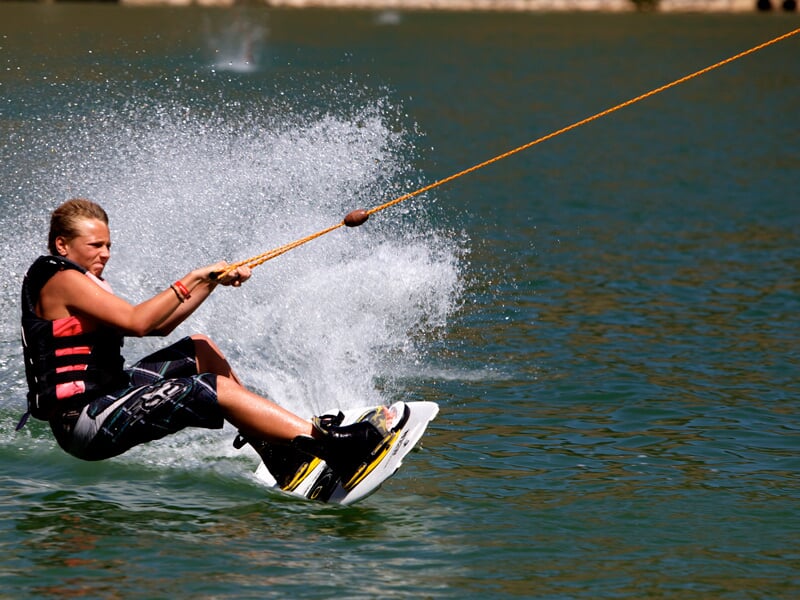 Une personne sur un wakeboard en équilibre sur son wakeboard, tirée par un câble de traction orange, créant un éclaboussement d'eau tout en glissant sur un lac calme