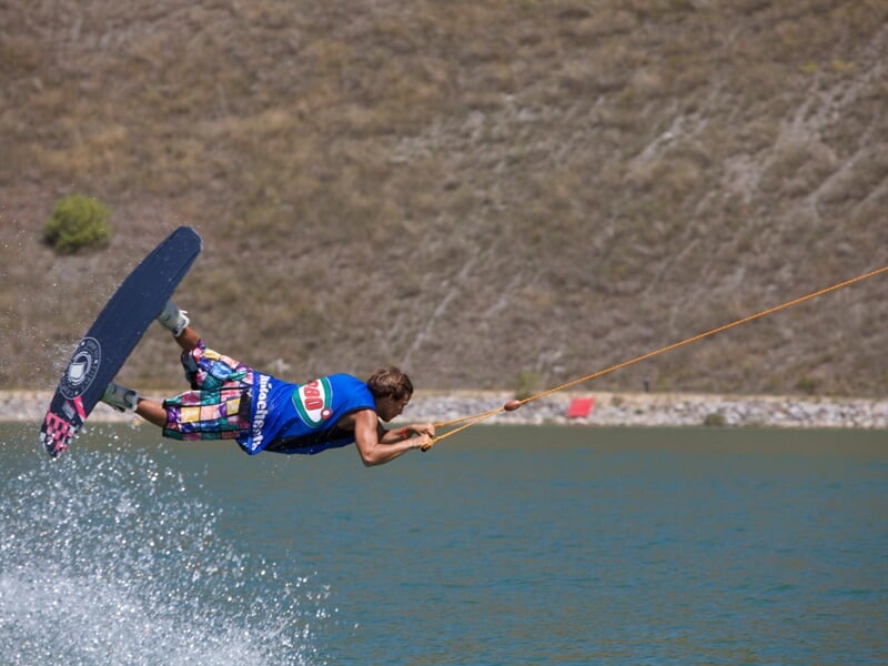 Une personne pratiquant le wakeboard en pleine action, effectuant un saut acrobatique élevé au-dessus de l'eau, tenue par un câble de traction orange, avec une colline en arrière-plan