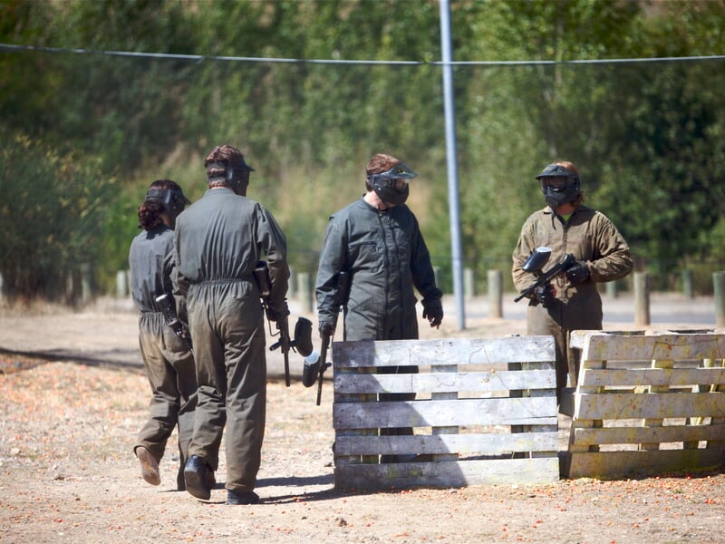 Quatre personnes en tenue de protection, avec des casques et des masques, marchent à côté de barrières en bois sur un terrain de jeu de paintball