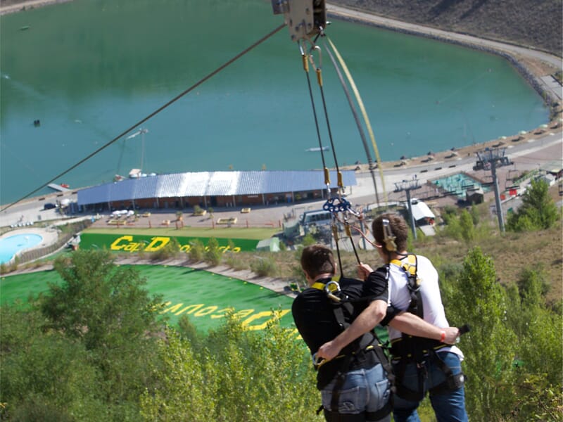 Deux personnes attachées à une tyrolienne au-dessus d'un lac et d'un espace de loisirs avec des installations et des bâtiments en contrebas