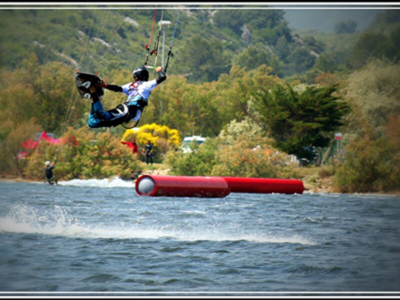 Kitesurfeur en combinaison bleue effectuant un saut au-dessus de l'eau avec un obstacle gonflable rouge en arrière-plan.