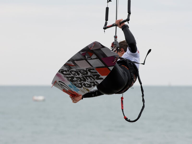 Kitesurfeur en combinaison noire et blanche réalisant une figure aérienne avec une planche colorée sur fond de mer.
