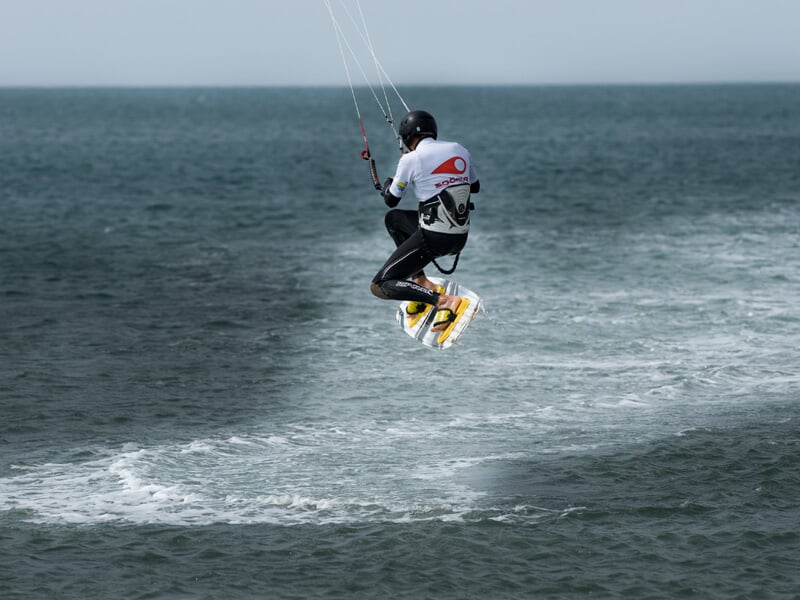 Kitesurfeur suspendu au-dessus de l'eau avec une combinaison blanche et noire, sur fond de mer agitée et ciel gris.