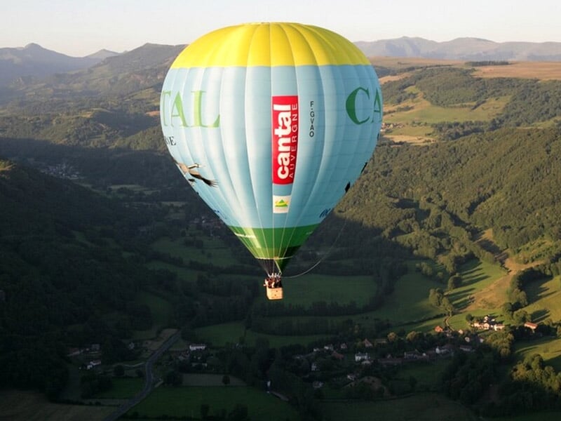 Montgolfière colorée avec inscription CANTAL survolant un paysage vallonné et boisé.