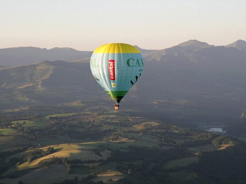 Montgolfière bleue et jaune avec inscription CANTAL survolant une région montagneuse au coucher du soleil.