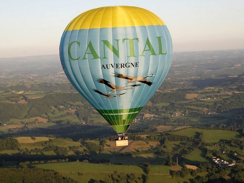 Montgolfière bleue avec inscription CANTAL et dessin d'oiseaux, survolant une campagne verte et vallonnée.