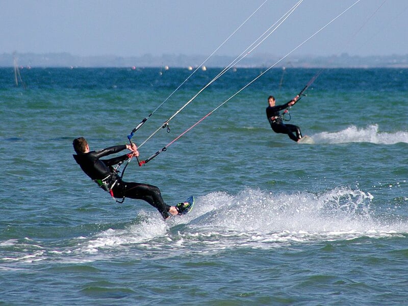 Deux kitesurfeurs glissent sur l'eau avec leurs ailes, créant des éclaboussures sous un ciel clair.