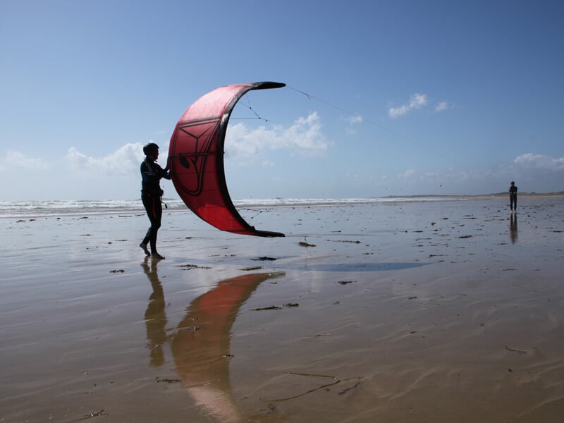 Personne tenant une aile de kitesurf rouge sur une plage mouillée, reflet visible sur le sable.