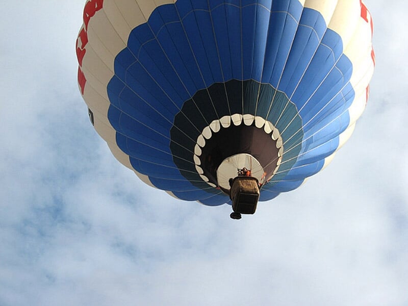 Montgolfière bleue vue de dessous, en vol dans un ciel partiellement nuageux, avec une nacelle suspendue.
