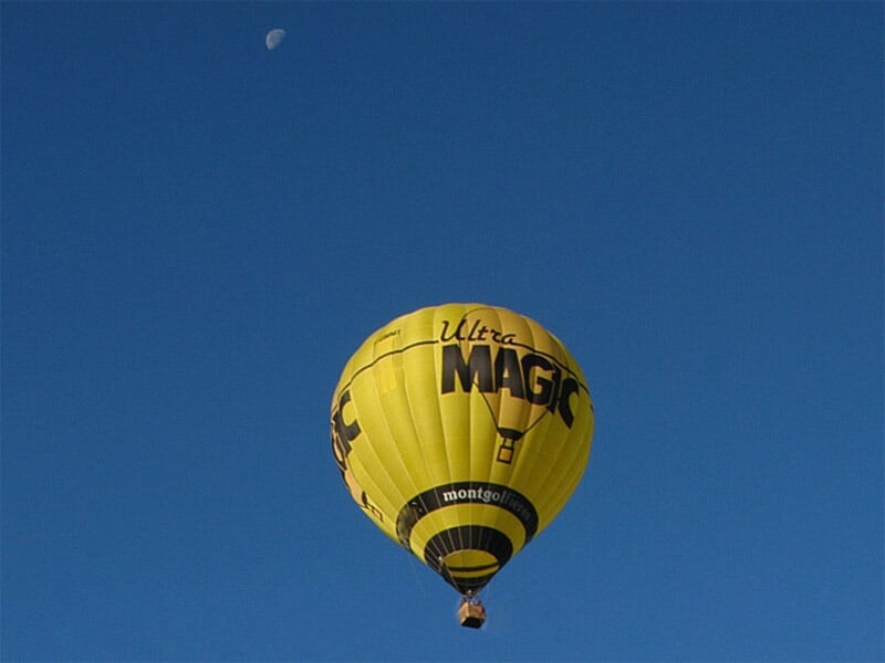 Montgolfière jaune avec inscription 'Ultra Magic' en vol dans un ciel bleu avec la lune visible.