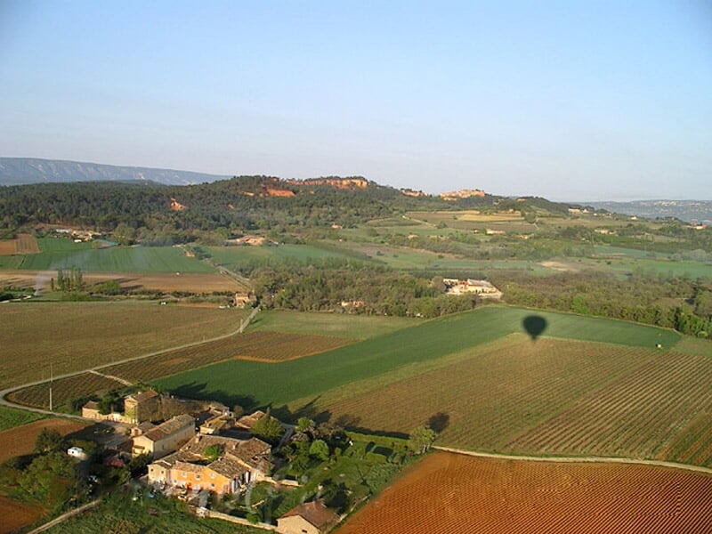 Vue aérienne d'un village entouré de champs verts et de collines, avec l'ombre d'une montgolfière au sol.