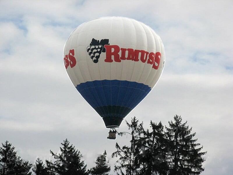 Montgolfière blanche et bleue avec logo 'Rimuss' en vol au-dessus d'une forêt dense et d'un ciel nuageux.