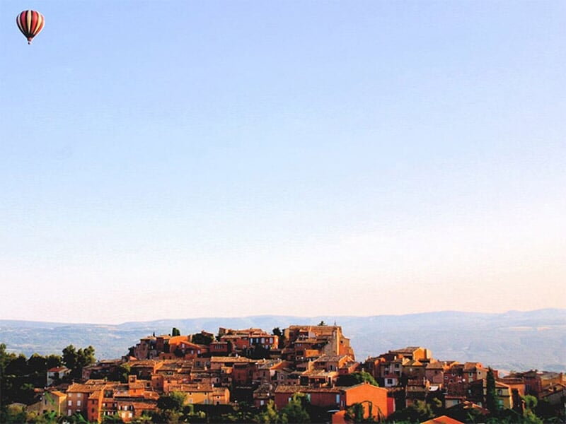 Montgolfière rouge et blanche en vol au-dessus d'un village perché sur une colline au coucher du soleil.