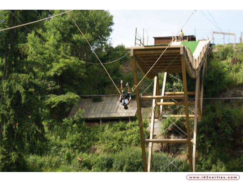 Plateforme en bois pour saut à l'élastique, personne suspendue tête en bas au-dessus d'un paysage verdoyant avec arbres et toiture en arrière-plan.