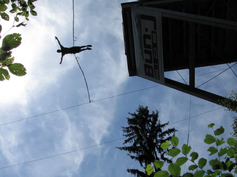 Saut en élastique depuis une tour élevée avec une silhouette humaine en suspension dans les airs, ciel partiellement nuageux et feuillage en contrebas, capturant l'adrénaline des sports extrêmes.