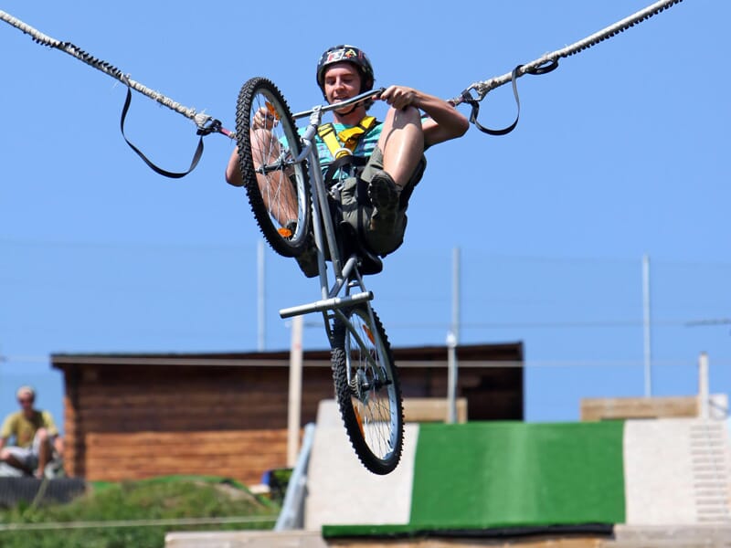 Cycliste suspendu sur un câble dans une activité d'aventure, avec un vélo en équilibre et un harnais de sécurité, ciel bleu dégagé et structure en bois en arrière-plan, mettant en avant le tourisme d'