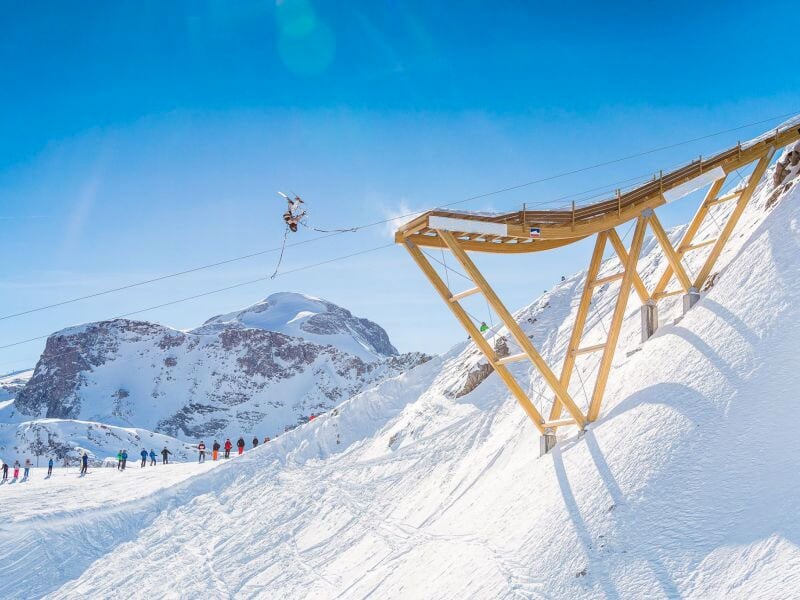 Vue large d'un saut à l'élastique en ski, capturé en plein air au-dessus d'une pente enneigée, avec des spectateurs en bas et des montagnes en arrière-plan