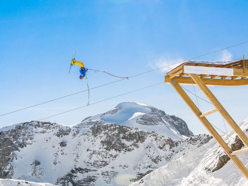 Skieur effectuant un saut périlleux à l'élastique depuis une structure en bois, au-dessus d'un paysage montagneux enneigé