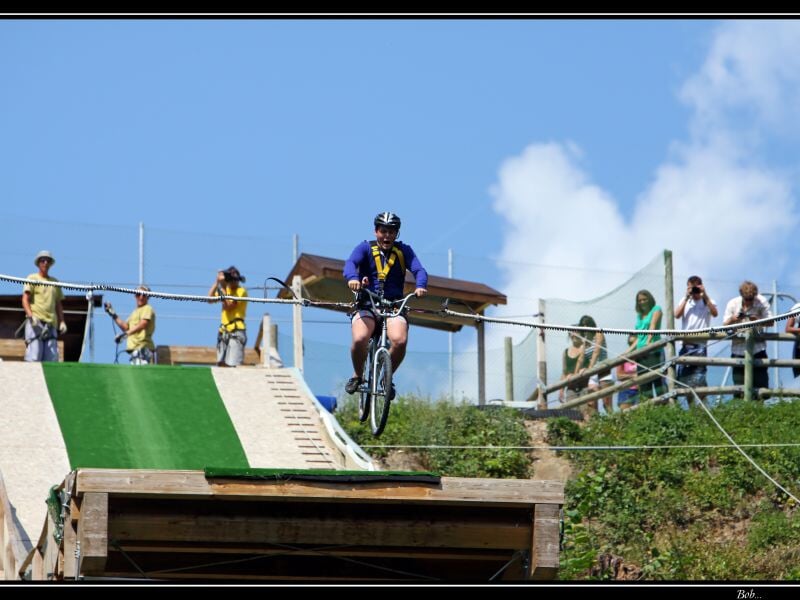 Participant roulant sur une plateforme étroite en vélo suspendu, entouré de spectateurs et personnel de sécurité, avec une rampe verte et un ciel dégagé.