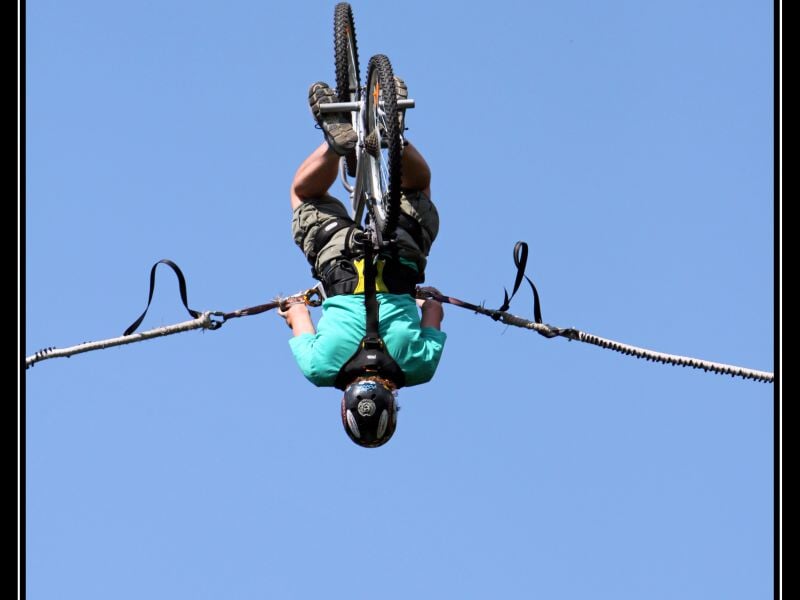 Cycliste suspendu la tête en bas lors d'une activité acrobatique en vélo sur filin, équipé d'un casque et harnais de sécurité, ciel bleu en arrière-plan.
