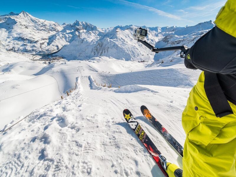 Vue en plongée d'un skieur équipé d'une caméra embarquée se préparant à descendre une pente enneigée, avec une chaîne de montagnes majestueuses en arrière-plan