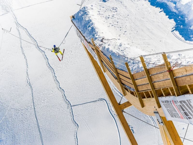Saut à l'élastique en ski depuis une plateforme en bois, avec une vue plongeante sur une vaste étendue de neige immaculée, entourée de montagnes enneigées
