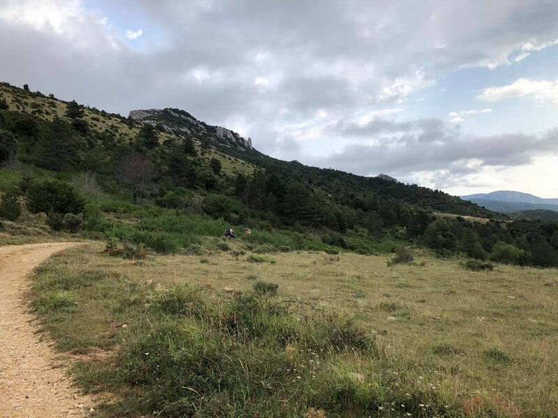 Chemin de campagne entouré de collines et de végétation dense sous un ciel partiellement nuageux.