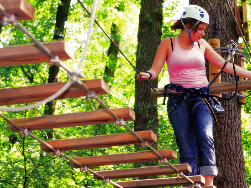Femme portant un casque et un harnais franchissant un pont de singe en bois dans un parcours aventure en forêt.