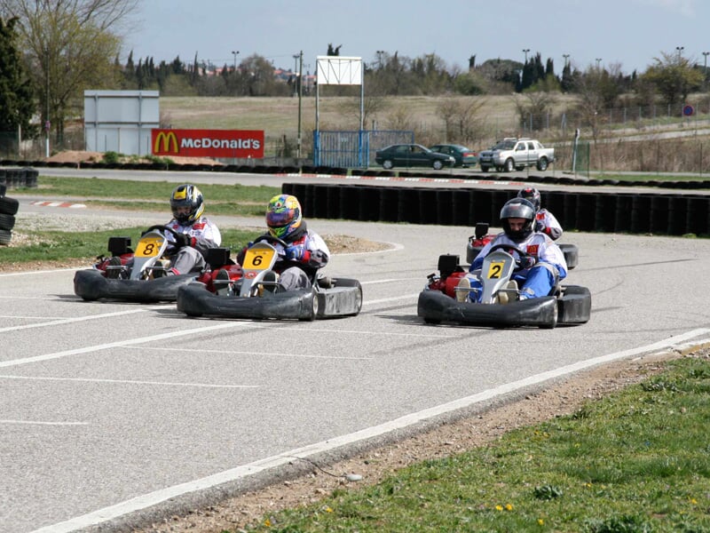 Trois pilotes en karting portant des casques colorés sur une piste extérieure avec un panneau McDonald's en arrière-plan.