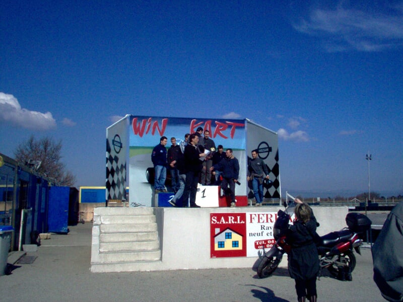 Podium de karting avec plusieurs personnes debout, un photographe prend une photo, ciel bleu et bâtiment en arrière-plan.