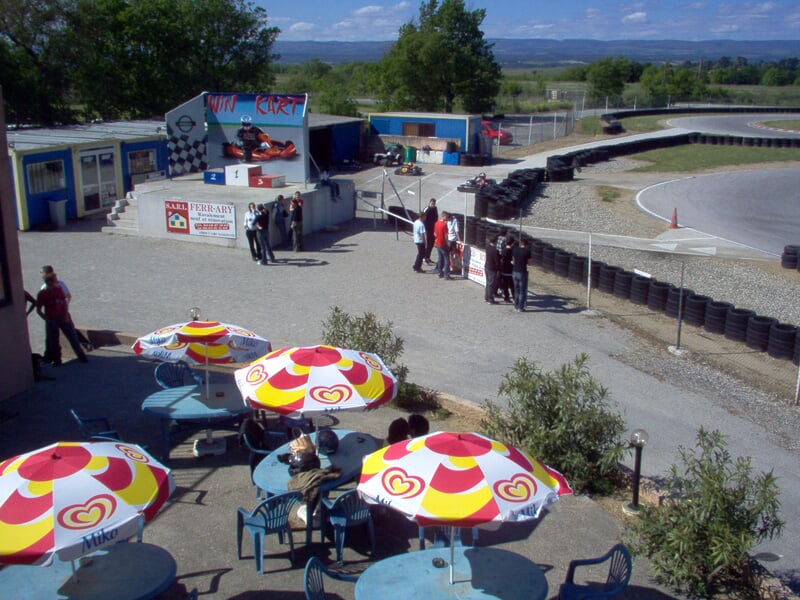 Vue aérienne d'une piste de karting avec des parasols colorés, des tables et des personnes autour de la zone de repos.