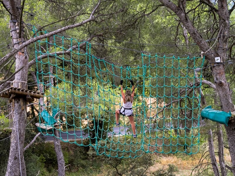 Une personne traverse un filet d'escalade suspendu entre les arbres dans un parc d'accrobranche.