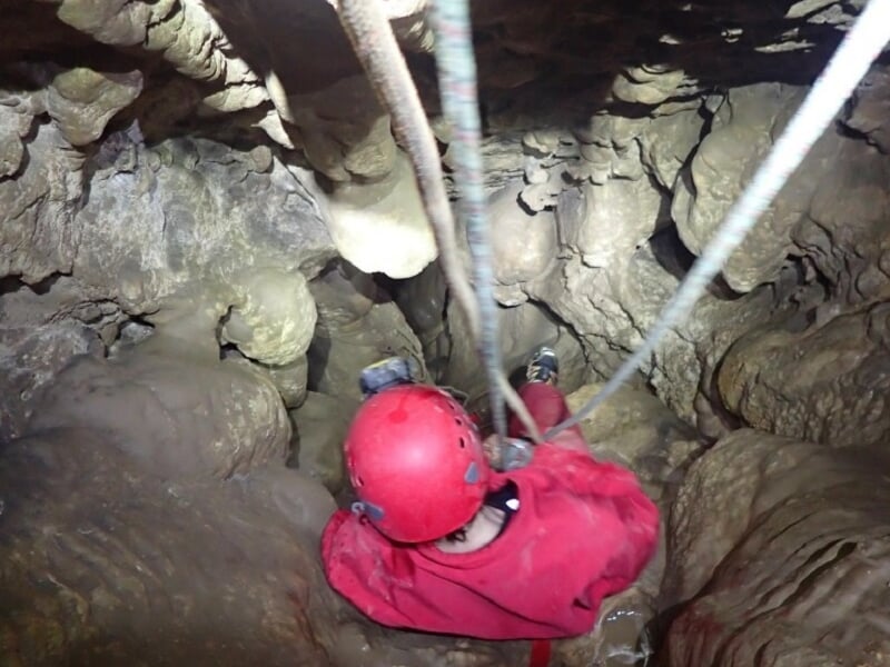 Spéléologue en combinaison rouge descend en rappel dans une cavité rocheuse sombre avec un casque et une lampe.