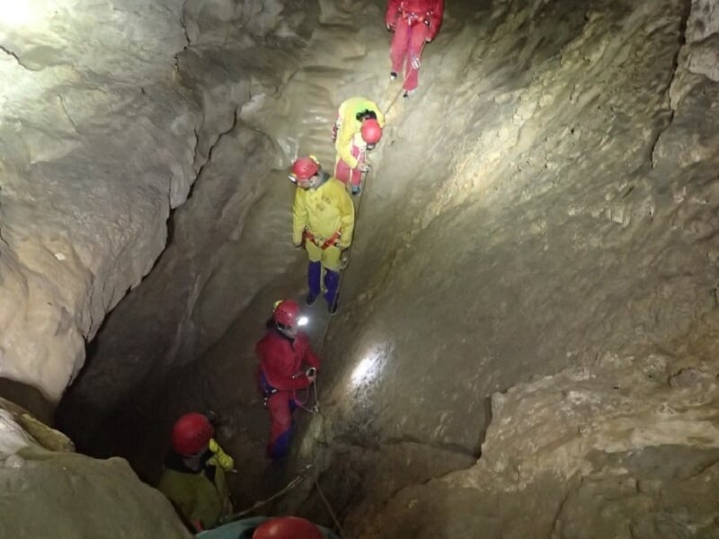 Plusieurs spéléologues progressent en file indienne dans une étroite galerie souterraine rocheuse éclairée.