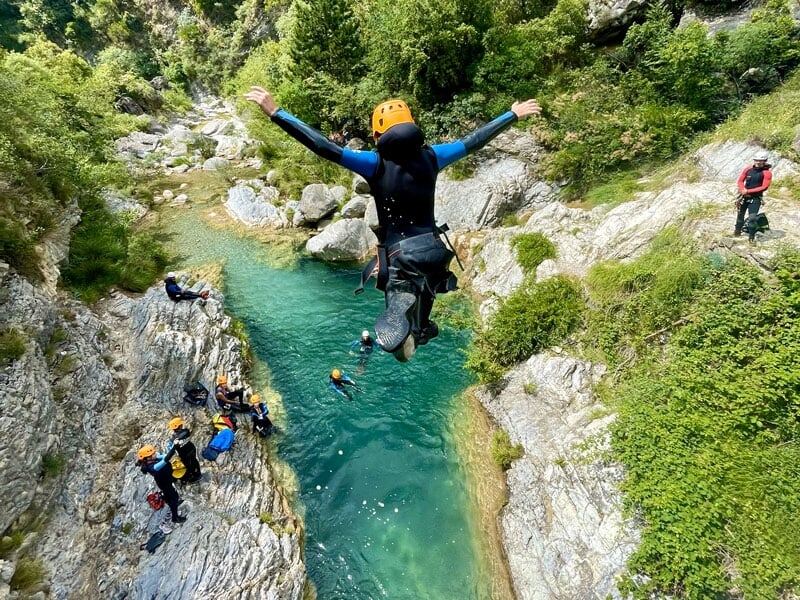 Saut d'une personne en combinaison noire et casque jaune, bras écartés, dans une eau turquoise au fond d'un canyon