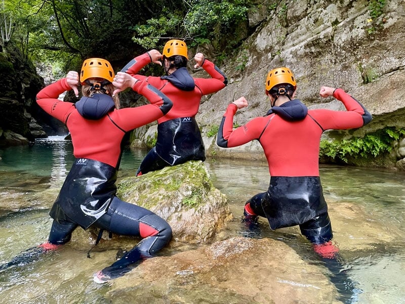Trois personnes en combinaison de canyoning, casques jaunes, posent dos à la caméra, montrant leurs muscles, assises sur des rochers dans une rivière peu profonde
