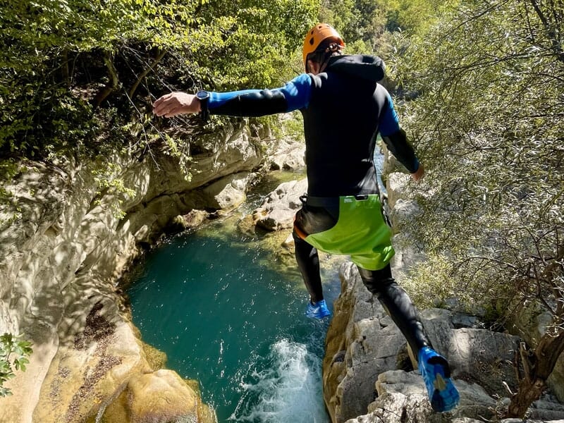 Saut d'une personne en combinaison de canyoning noir et bleu, au-dessus d'une rivière entourée de parois rocheuses, sous un ciel ensoleillé