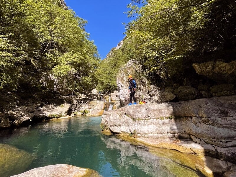Vue d'un canyon avec un homme équipé de matériel de canyoning, debout sur un rocher au bord d'une rivière calme et turquoise
