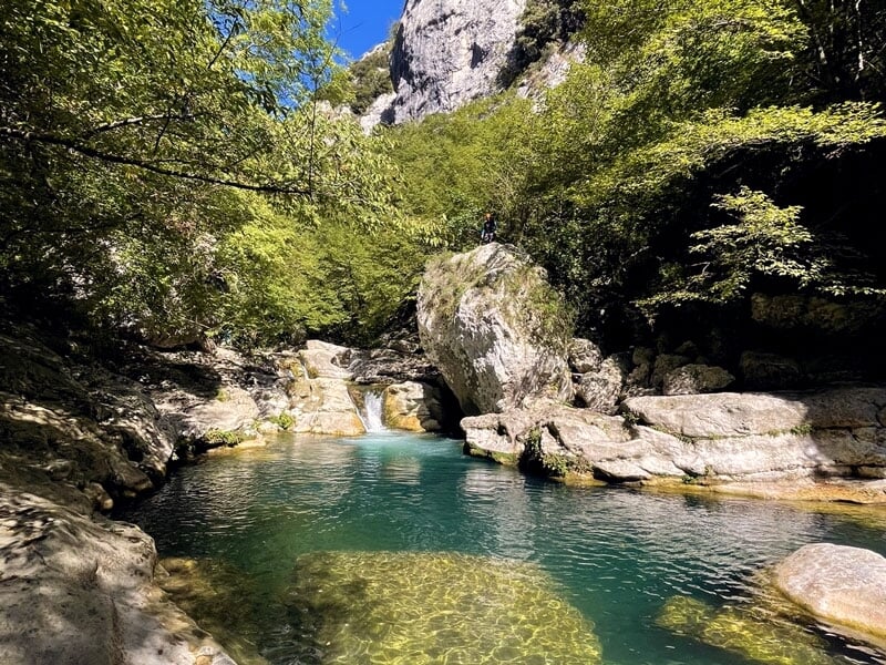 Paysage de canyon avec eau turquoise entourée de rochers et de végétation dense, sous un ciel bleu clair