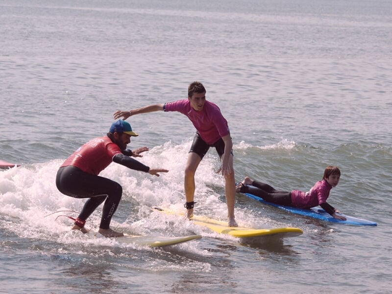 Trois surfeurs débutants portant des combinaisons roses et noires, apprenant à surfer sur de petites vagues à Longeville-sur-Mer.